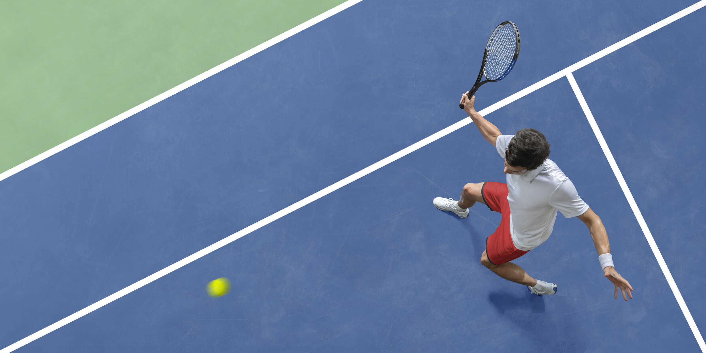 overhead view of a male tennis player in white shirt and red shorts, holding up a racket in mid action, about to strike the ball in a volley during a tennis match