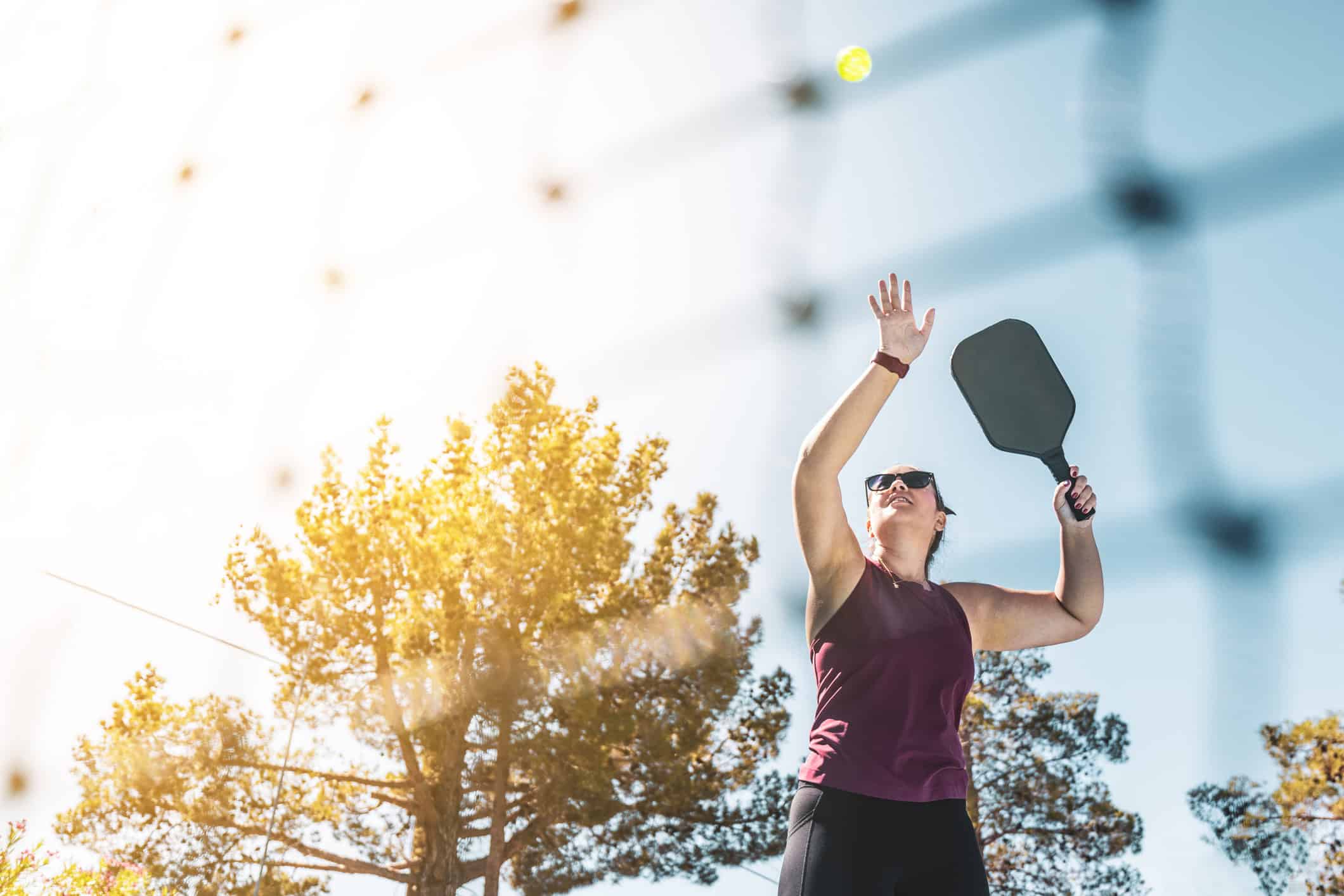 woman hitting a pickleball in the air above her ahead on a beautiful sunny day with trees in background