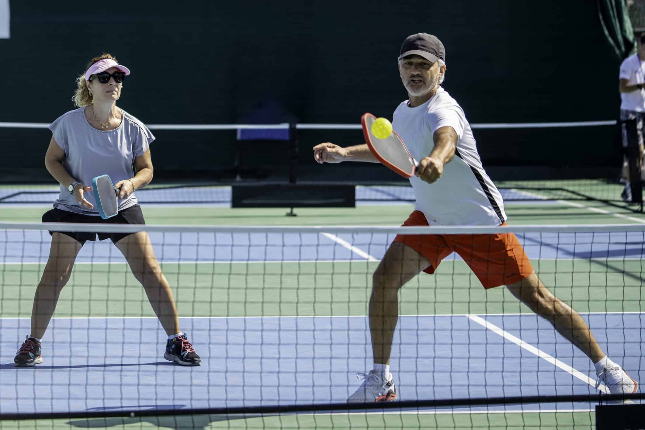 male and female pickleball players are playing, male player is shooting forehand with ball on racket, female pickleball player is on return horizontal